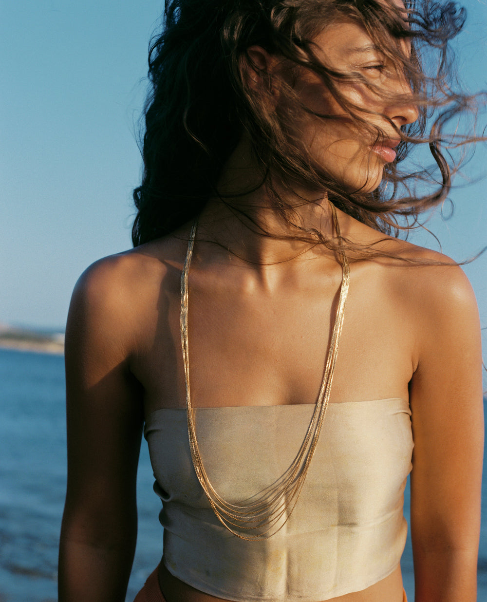 A woman outdoors by the water wearing a wide, strapless cream top and a long layered gold necklace, with her hair blowing in the wind against a blue sky.