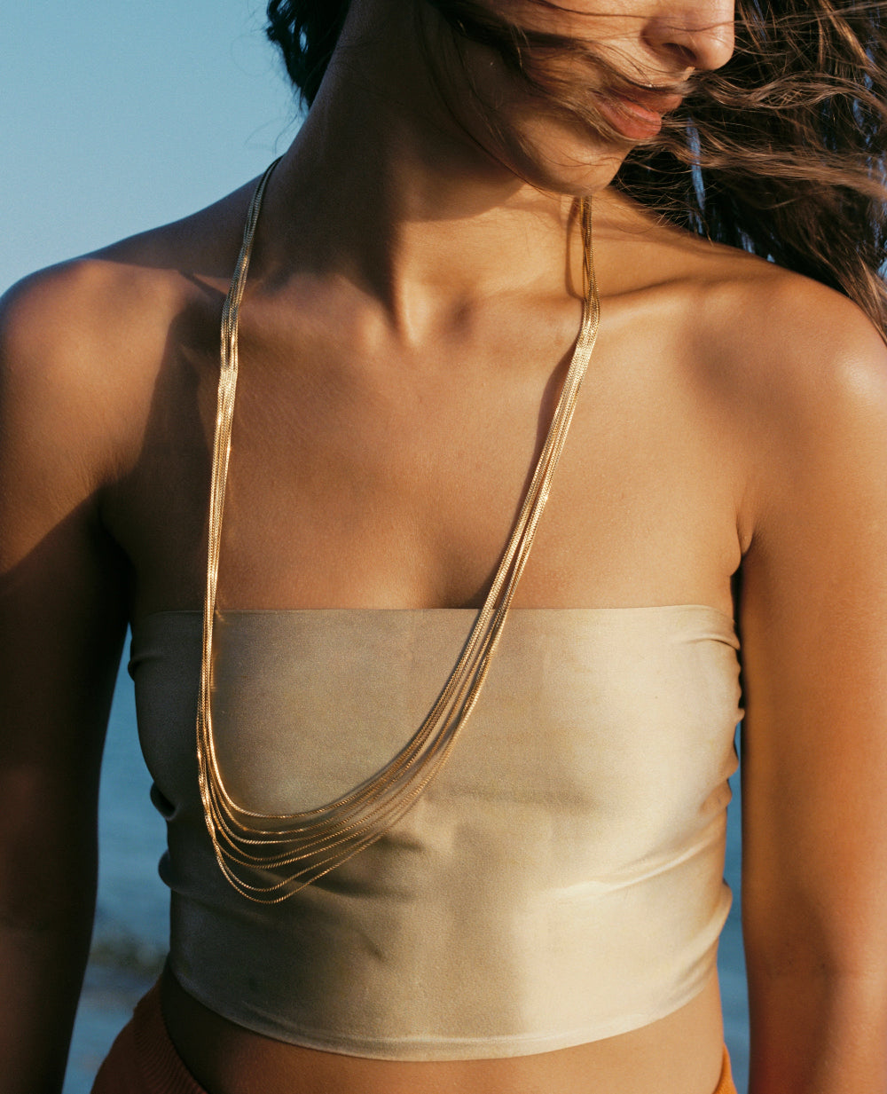 A close-up lifestyle shot of a model wearing a multi-strand gold chain necklace over a beige satin bandeau top, set against a soft blue background.